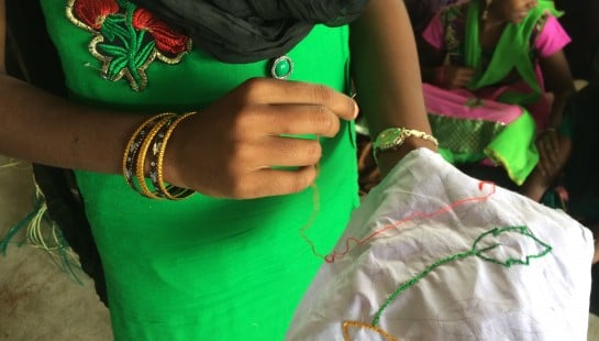 An image of an Indian girl in a green dress, sewing a piece of fabric