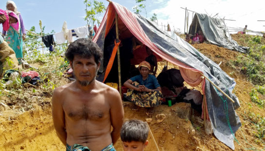 A Rohingyan refugee stands with his son in front of their emergency shelter