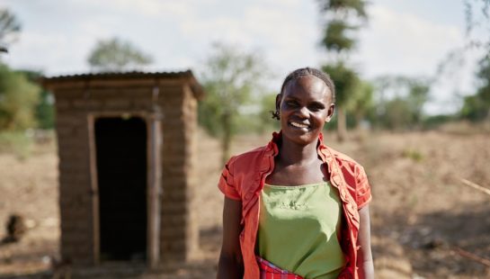 A woman smiles at the camera in front of her outhouse