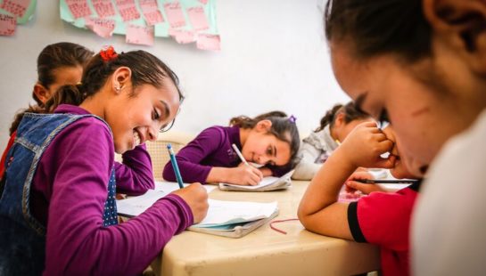 Children sit at a desk writing in their notebooks