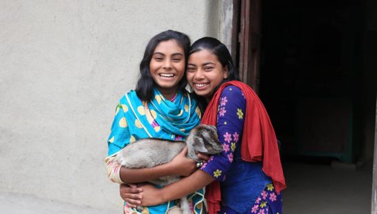 Two girls with a goat stand in front of a building in Bangladesh.