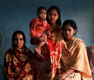 Three Bangladeshi women of varying ages and a baby stand and sit in a dark blue room.