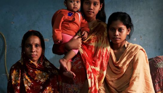 Three Bangladeshi women of varying ages and a baby stand and sit in a dark blue room.