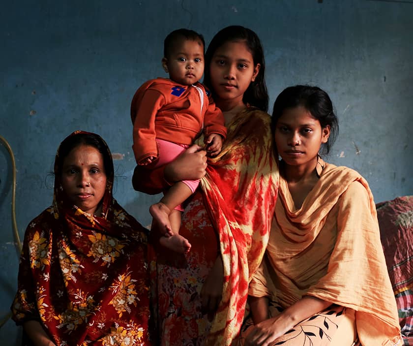 Three Bangladeshi women of varying ages and a baby stand and sit in a dark blue room.