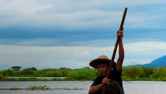 A man sits on the edge of a boat with an oar in hand