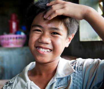 A young boy with a hand to his head smiles at the camera.