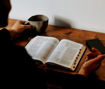 A man sits at a desk with an open Bible and a mug in hand