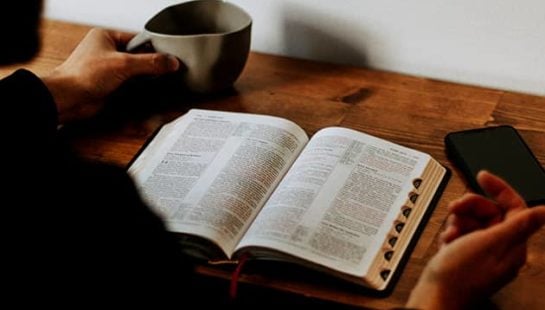 A man sits at a desk with an open Bible and a mug in hand
