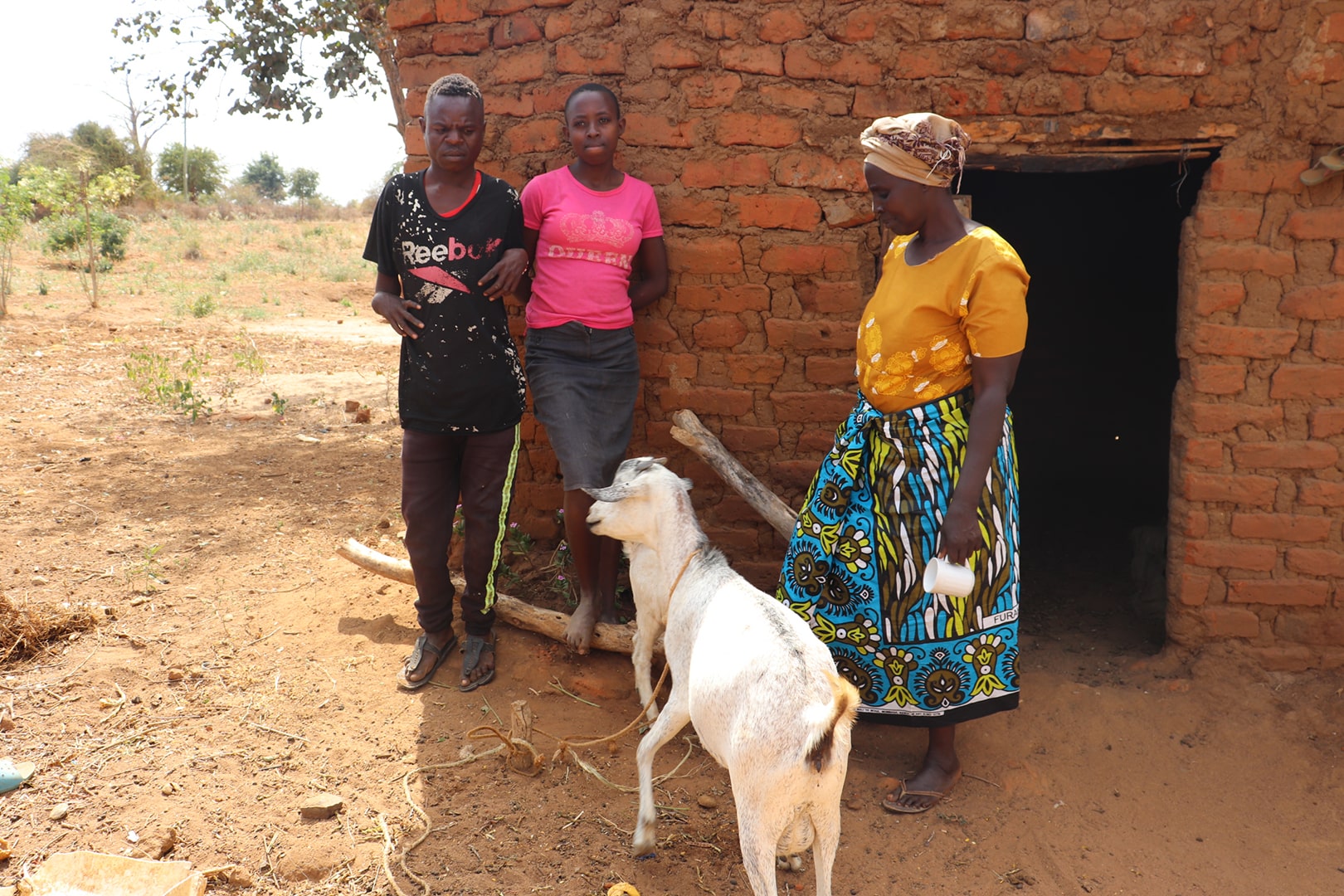 Three people stand in front of a brick hut with a goat