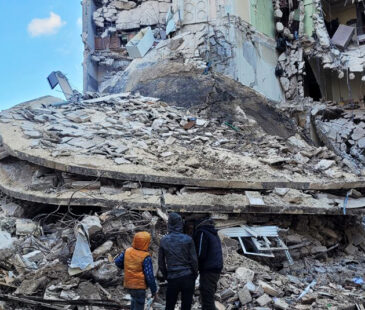 Three boys standing facing a building damaged by an earthquake