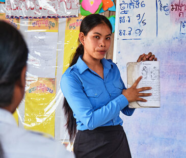 Cambodian teacher in a blue shirt stands in front of class holding a book